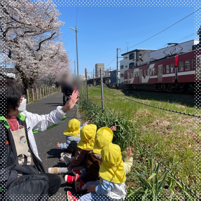 桜の下で電車に手をふる子どもと保育士