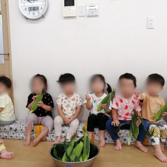 皮をむく前のとうもろこしと記念撮影する2歳児